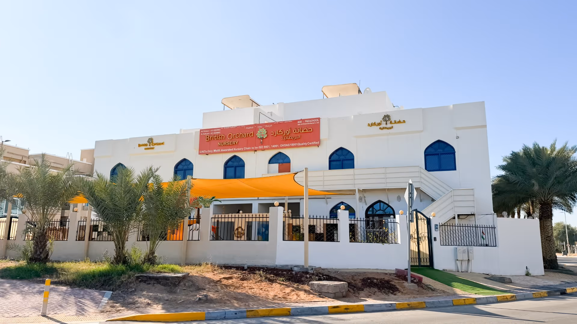 Two-story white building with blue arched windows and signs reading British Orchard Nursery, surrounded by palm trees and a black fence.