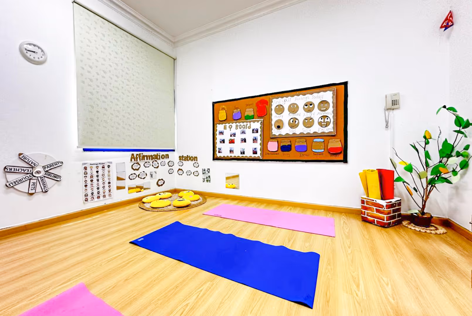 Bright classroom corner with wooden floor, colorful yoga mats, smiley face cushions, affirmation and feelings boards, and a decorative plant.