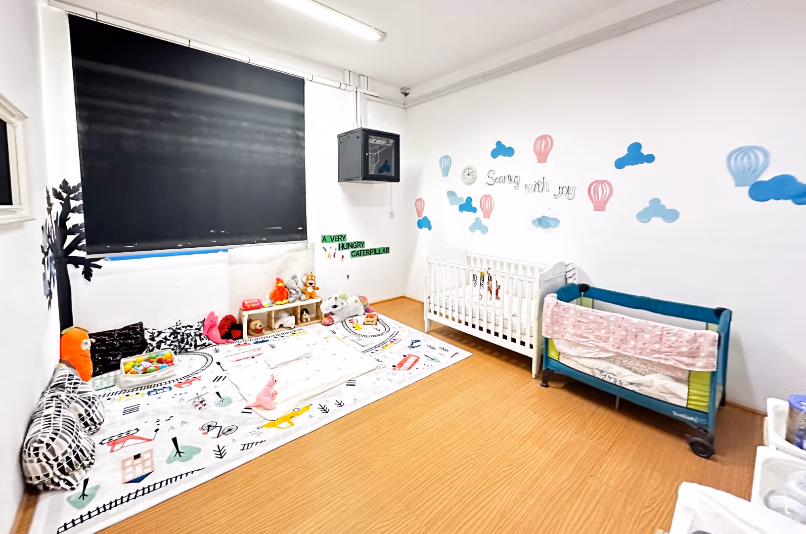 Bright nursery room with two cribs, a play mat with toys, stuffed animals, and wall decorations including clouds and hot air balloons.