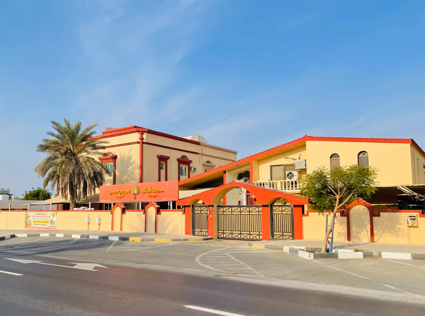 Exterior view of a yellow and red two-story building with a gated entrance, palm trees, and a clear blue sky.