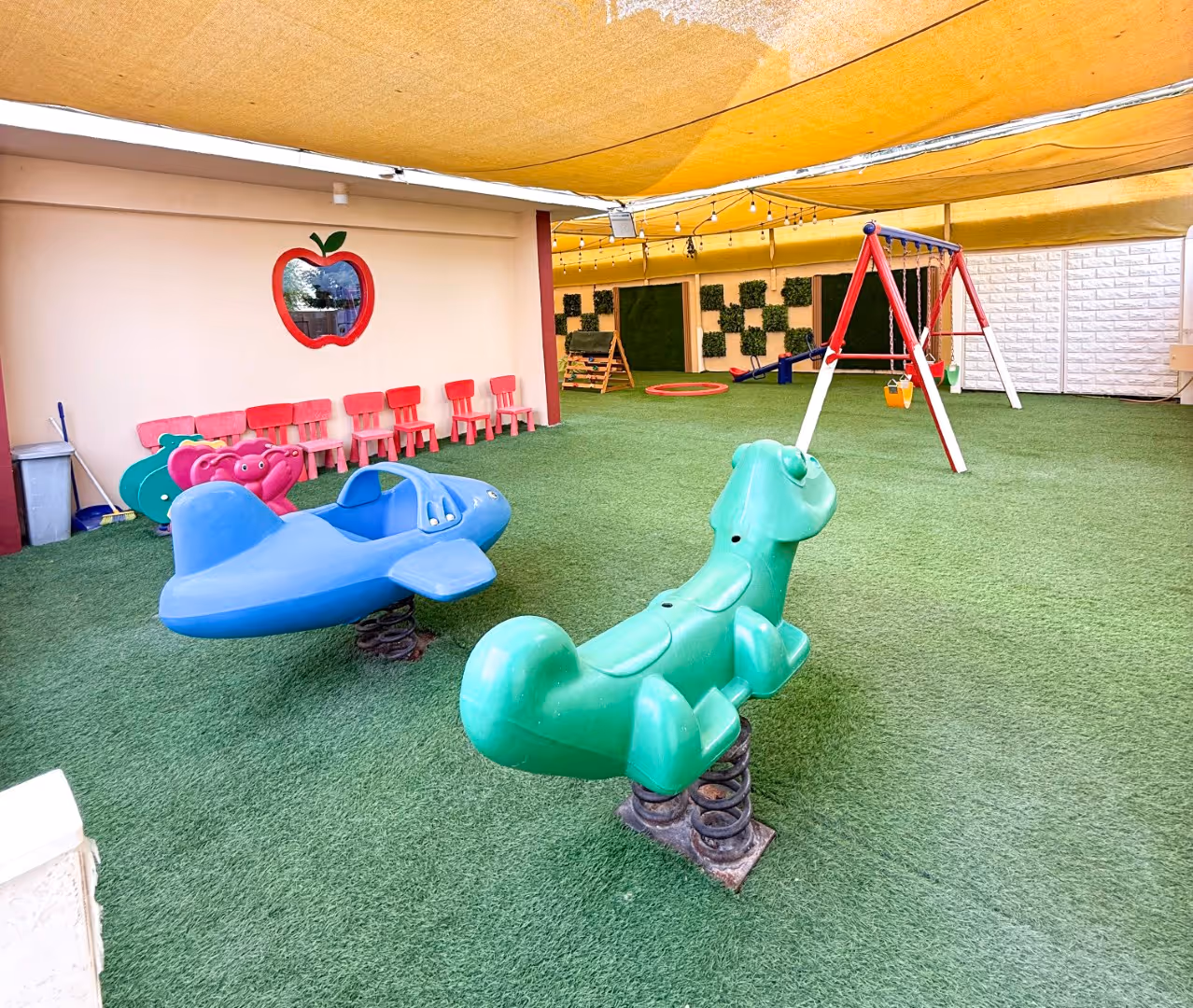 Indoor children's play area with green artificial grass, spring riders shaped like a green dog and blue airplane, red and green swings, and small red chairs lined against a wall with an apple-shaped mirror.