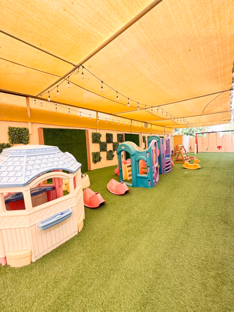 Covered playground area with artificial grass featuring plastic play houses, slides, swings, and a rocking duck.