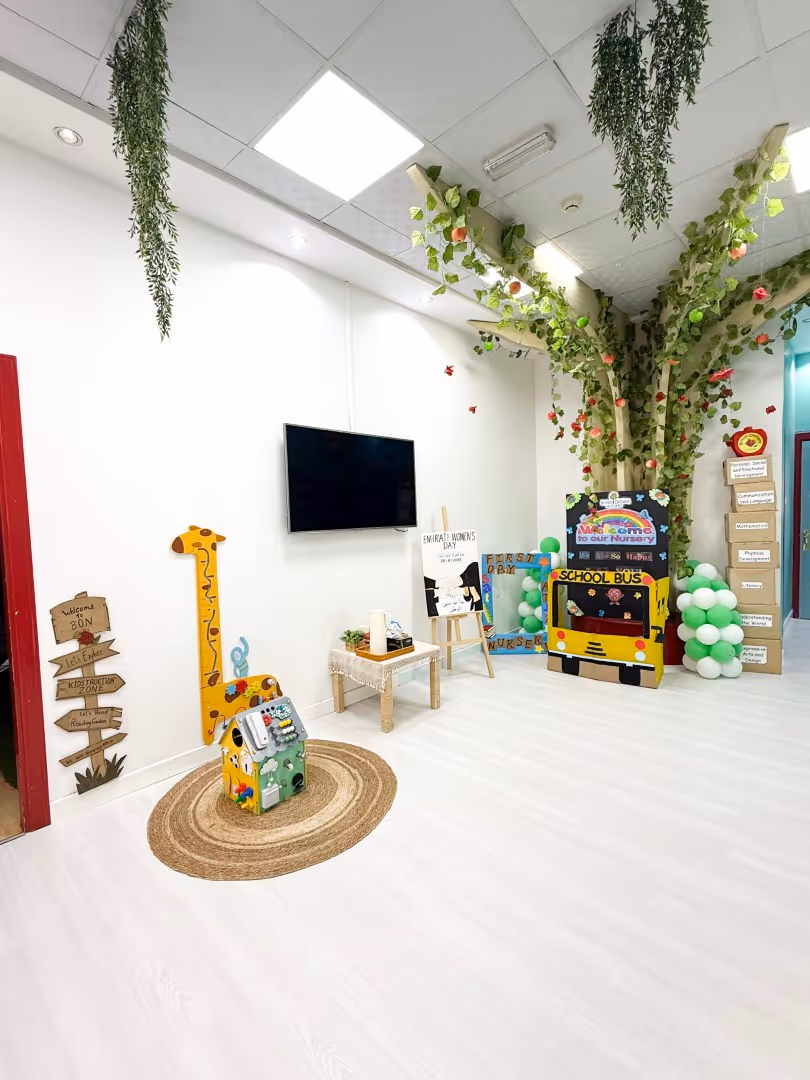 Bright nursery corner with a decorative tree, yellow school bus display, toy house, giraffe height chart, and a wall-mounted TV.
