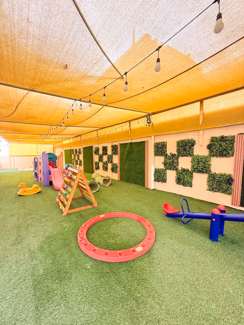 Covered outdoor children's play area with artificial grass, plastic slides, toy swings, tunnels, and climbing frame under string lights.