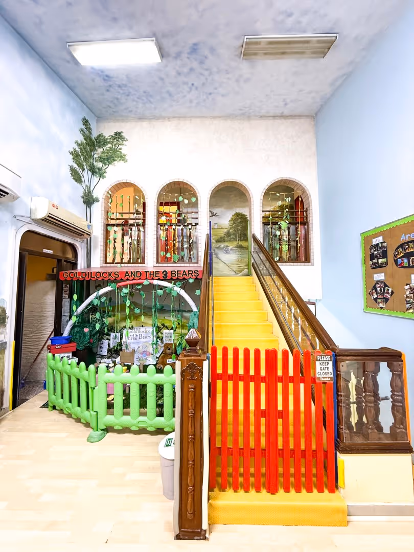 Indoor play area with yellow stairs blocked by a red gate, a green fenced section decorated with plants and signs reading 'Goldilocks and the 3 Bears.'