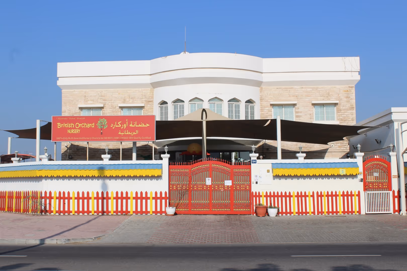 Facade of British Orchard Nursery with a white building, red decorative gate, and colorful fence under a clear blue sky.