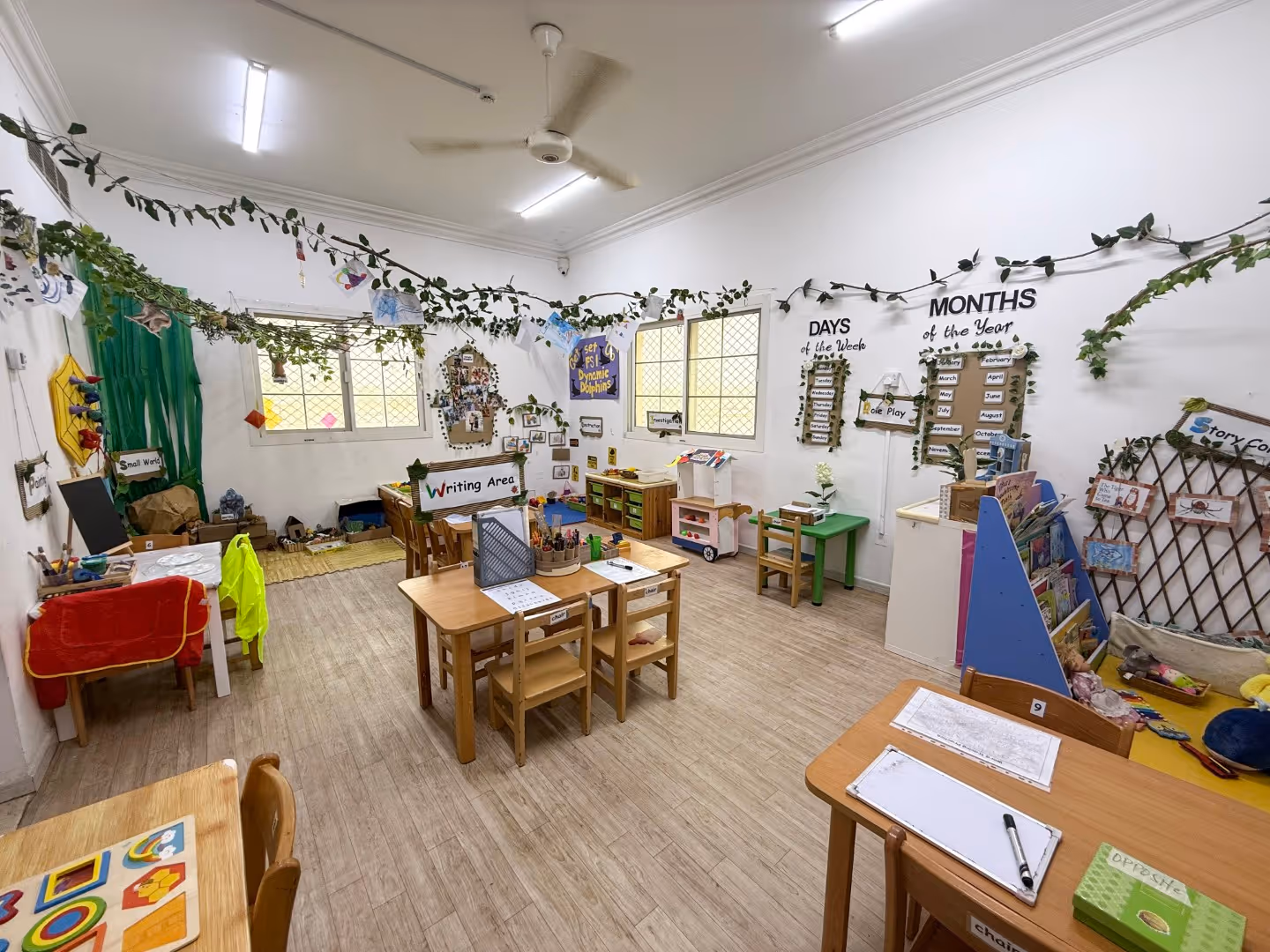 Bright and organized preschool classroom with small wooden tables and chairs, a writing area, educational posters for days and months on the wall, and creative decorations with greenery hanging from the ceiling.