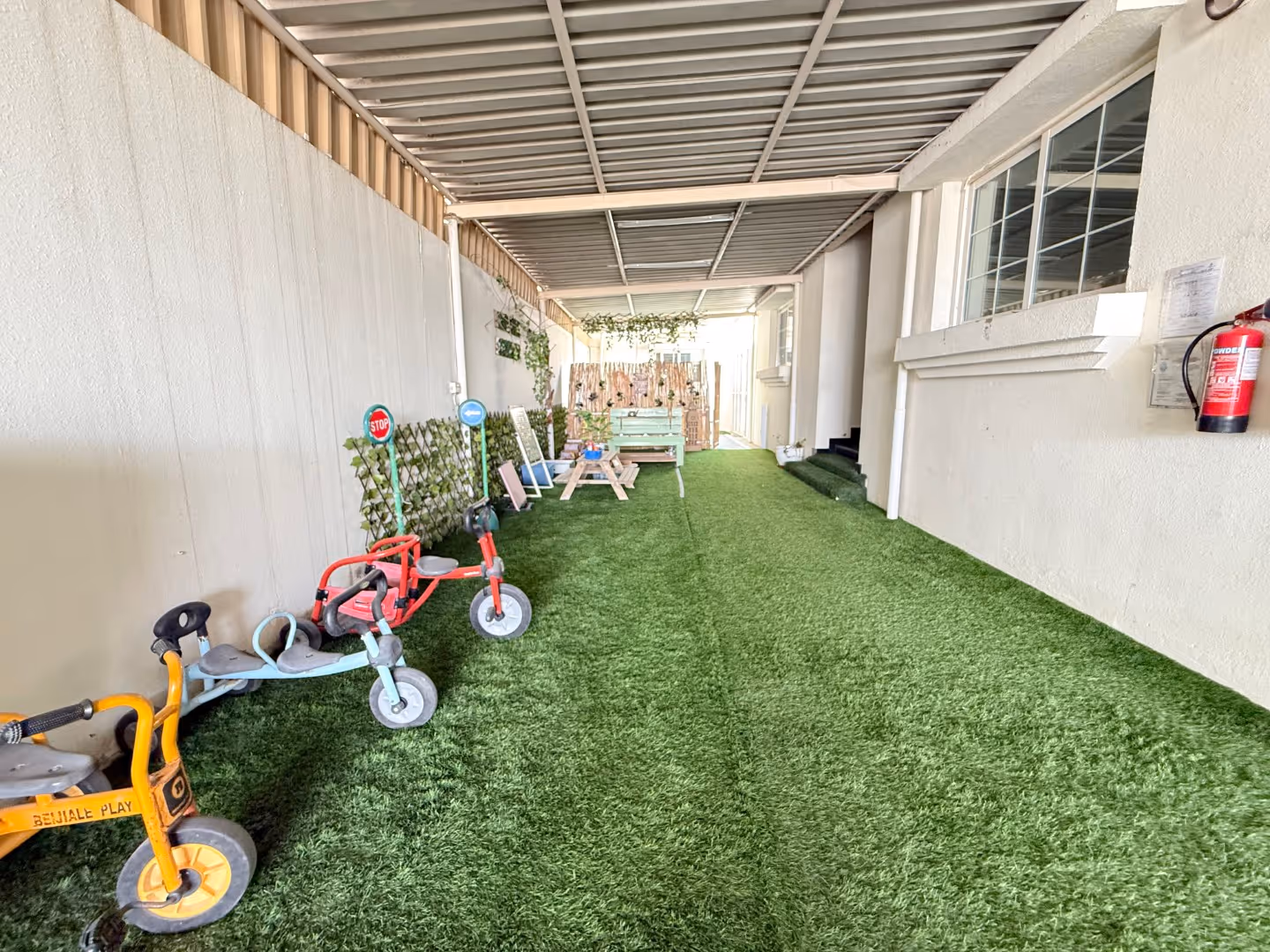 Covered play area with artificial grass, small tricycles, a picnic table, a bench, and wall-mounted plants.
