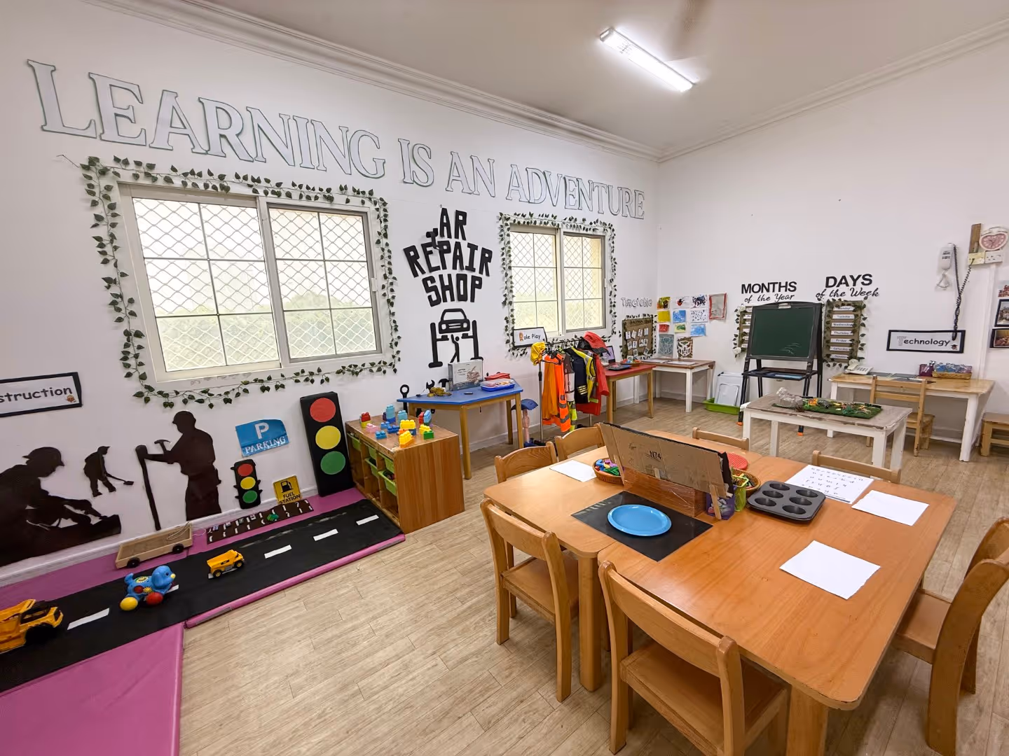 Classroom corner with wooden tables and chairs, educational play areas including a car repair shop theme, months and days charts, and a wall phrase saying Learning is an Adventure.