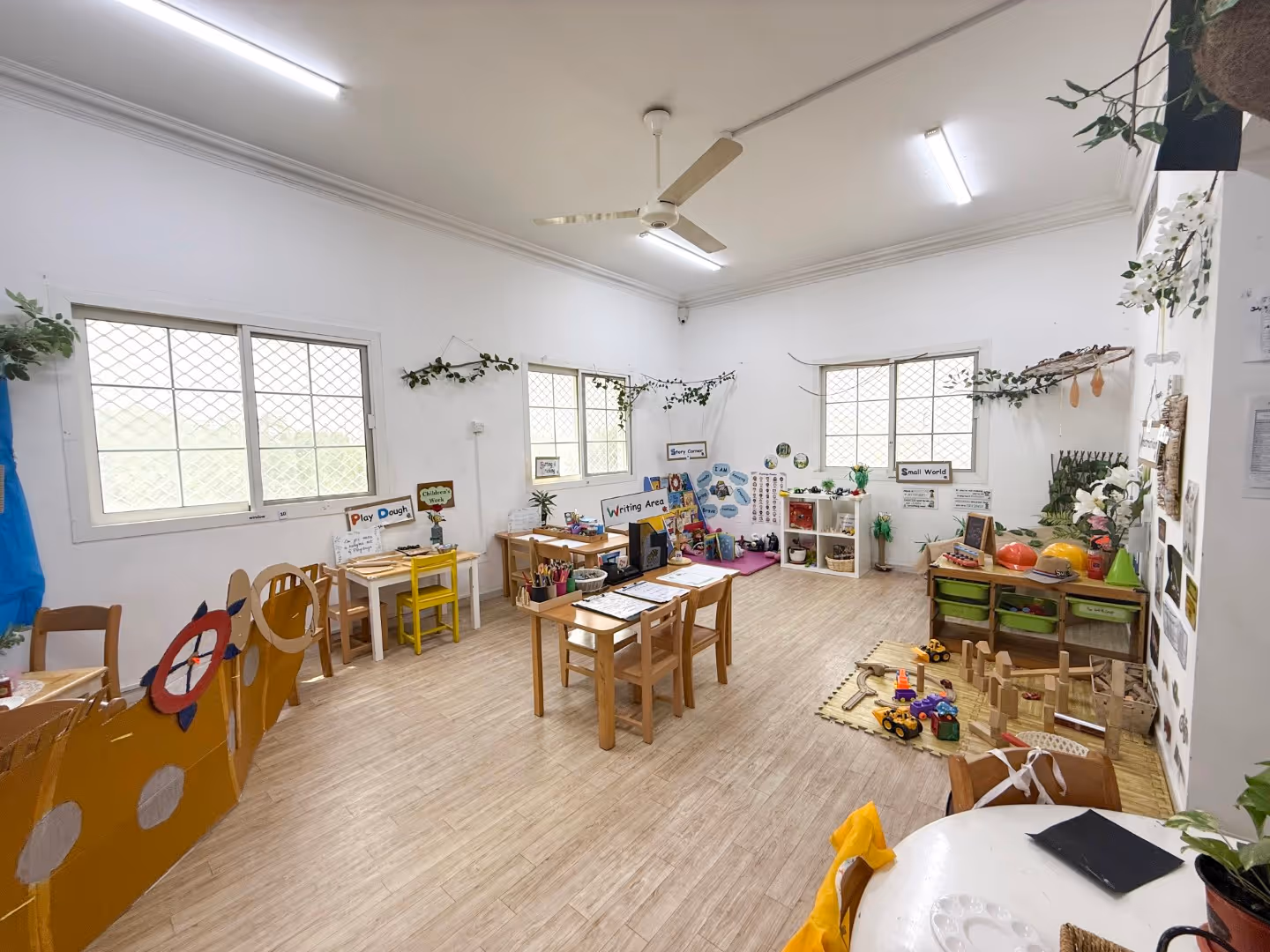 Bright and spacious classroom with wooden furniture, play areas, and educational toys under ceiling fans and fluorescent lights.