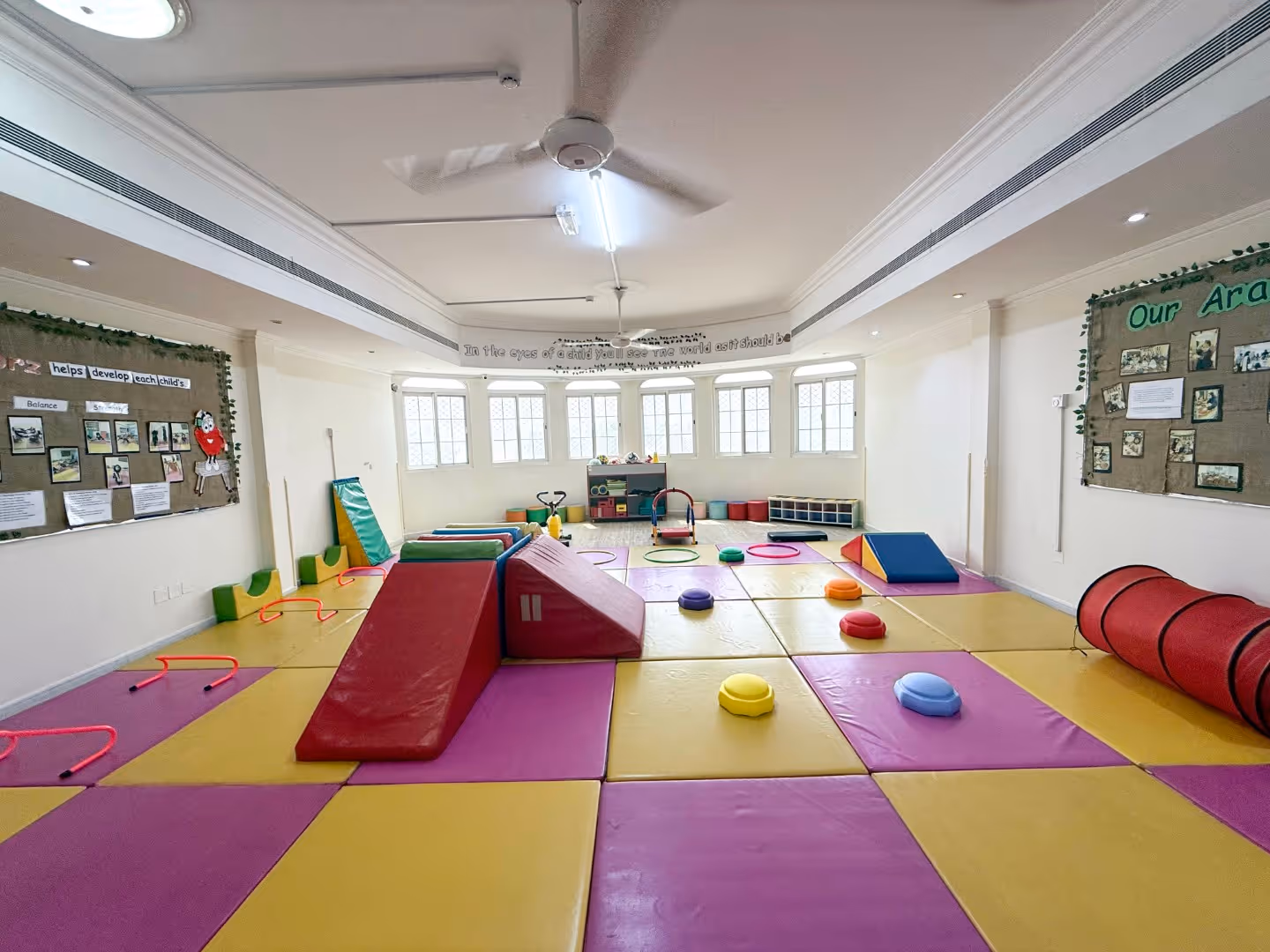 Bright indoor children's playroom with colorful padded floor mats, soft climbing ramps, small hurdles, play tunnels, and wall boards with photos and information.