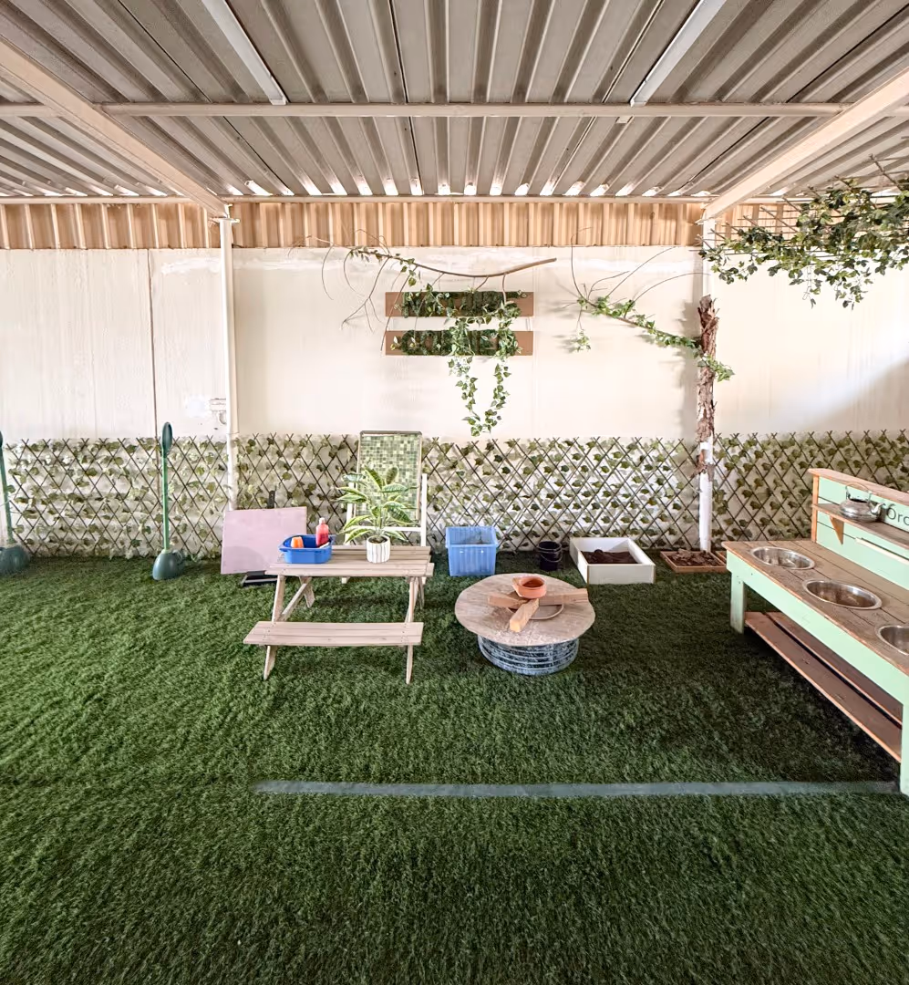 Indoor garden play area with artificial grass, a small wooden picnic table, a round wooden table, plants, and a mint-green play kitchen with metal sinks.