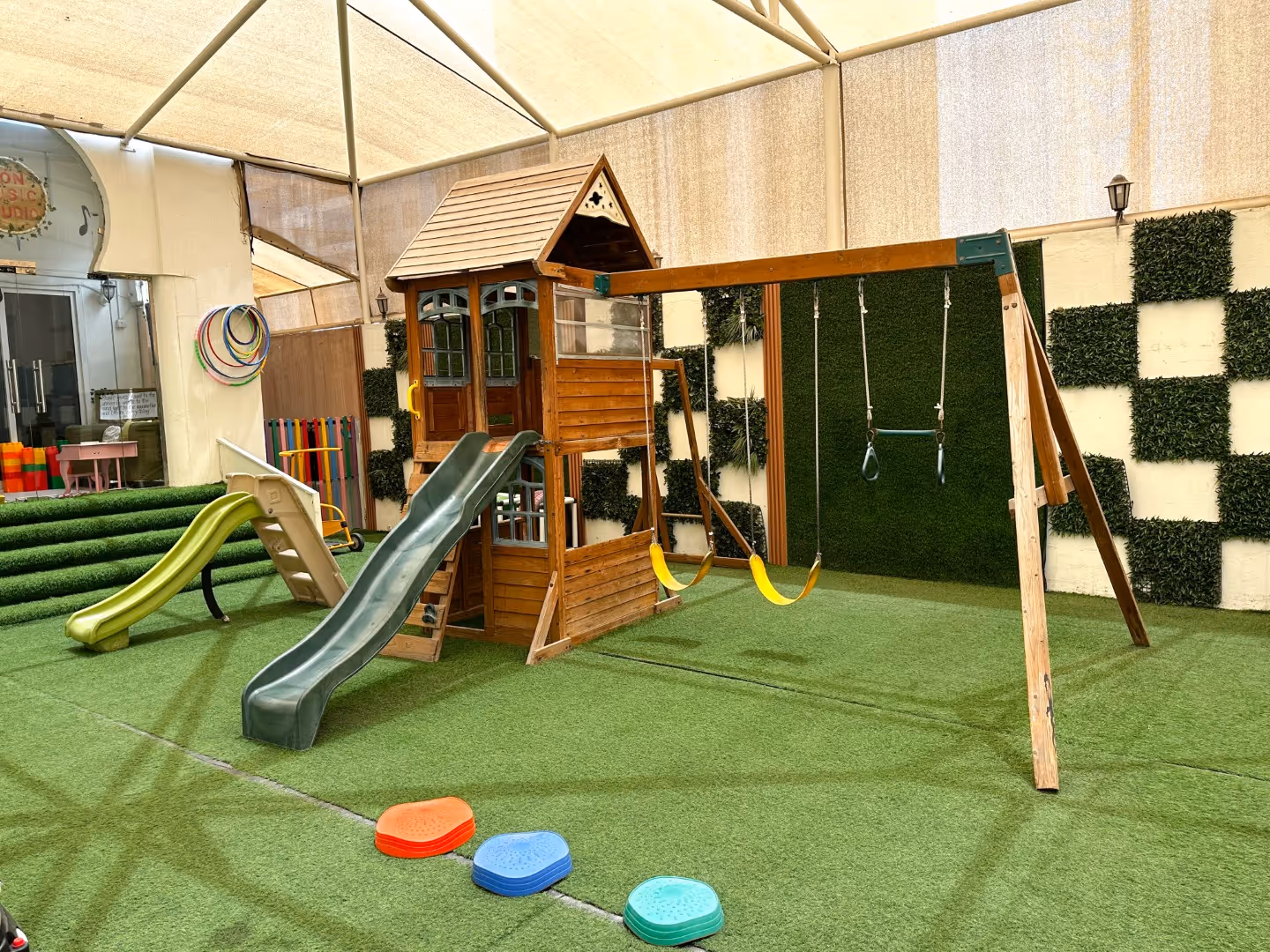 Indoor playground with wooden playhouse, two slides, two yellow swings, gymnastics rings, and artificial grass flooring.