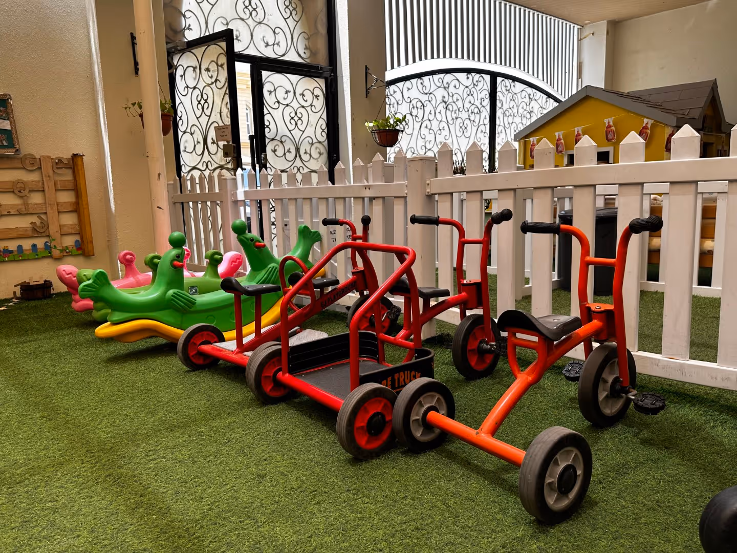 Children's indoor play area featuring green and pink animal-shaped rockers and red pedal vehicles on artificial grass next to a white picket fence.