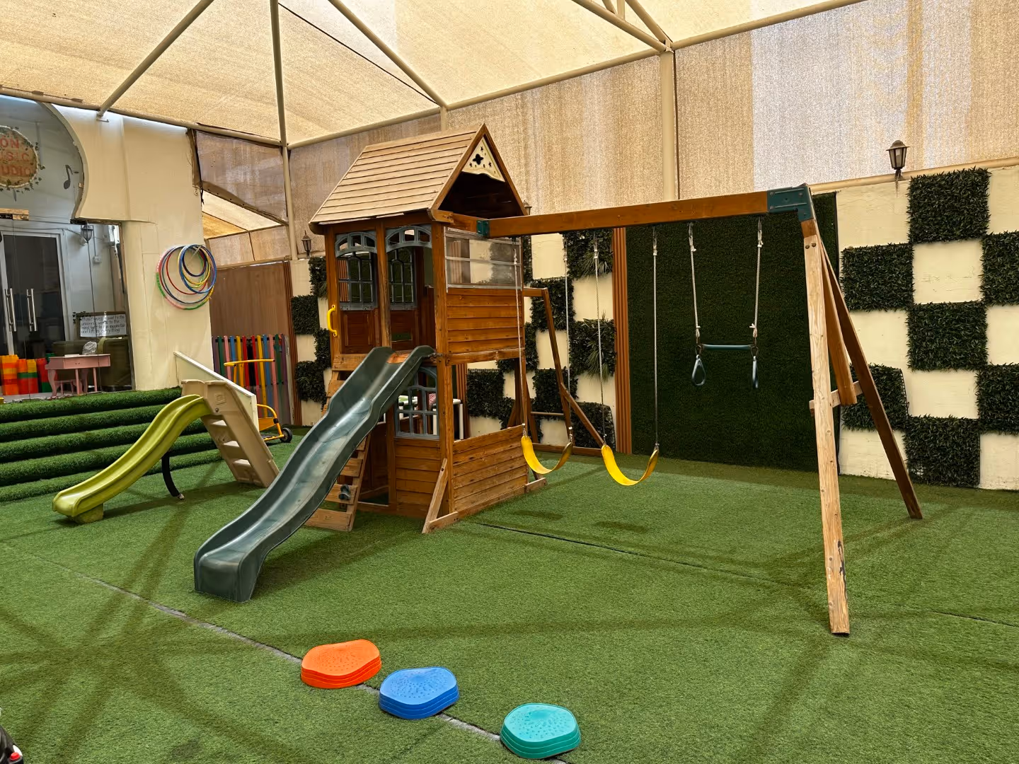 Indoor playground with artificial grass flooring featuring a wooden playhouse with slides and two yellow swings under a beige canopy.