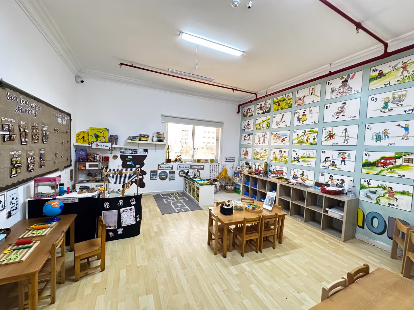Bright classroom with wooden tables, children's chairs, alphabet wall posters, toys, and learning stations.