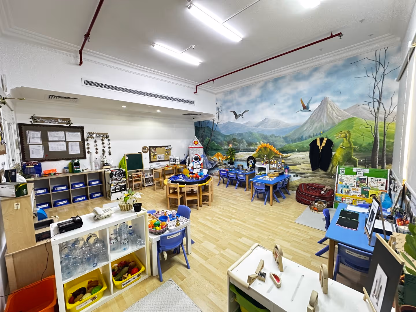 Colorful and organized children's classroom with dinosaur mural, tables, chairs, toys, and educational materials.