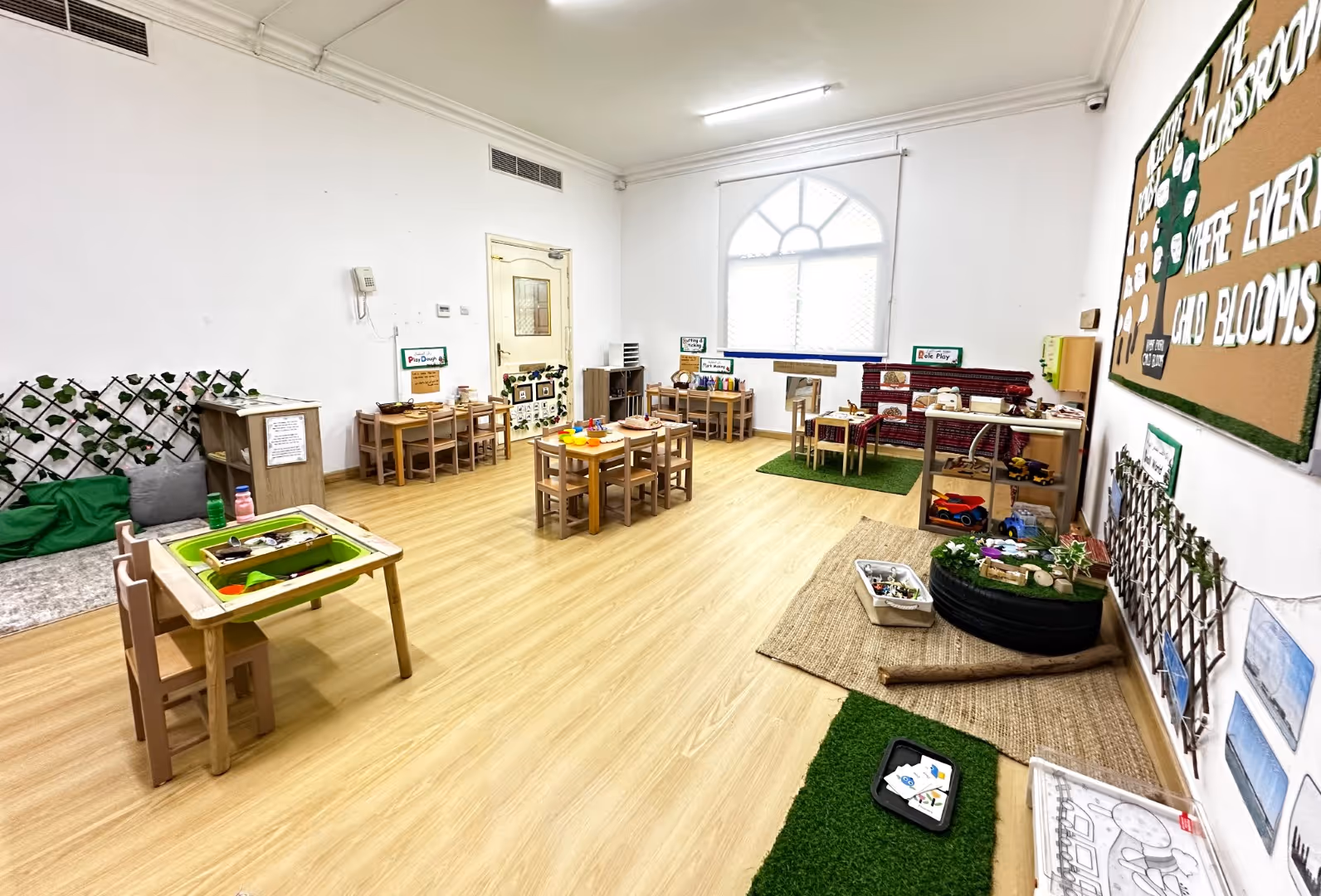 Bright and organized early childhood classroom with small wooden tables and chairs, educational toys, and learning stations.