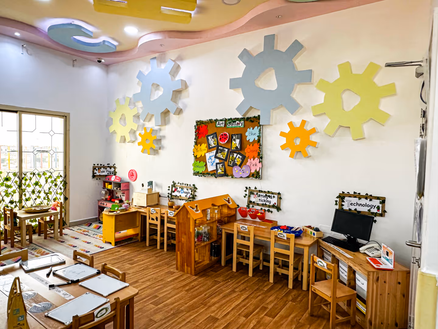 Colorful children's classroom with wooden furniture, tables, small chairs, educational play areas labeled role play, play dough, cutting & sticking, and technology, decorated with large gear-shaped wall art and a corkboard art gallery.