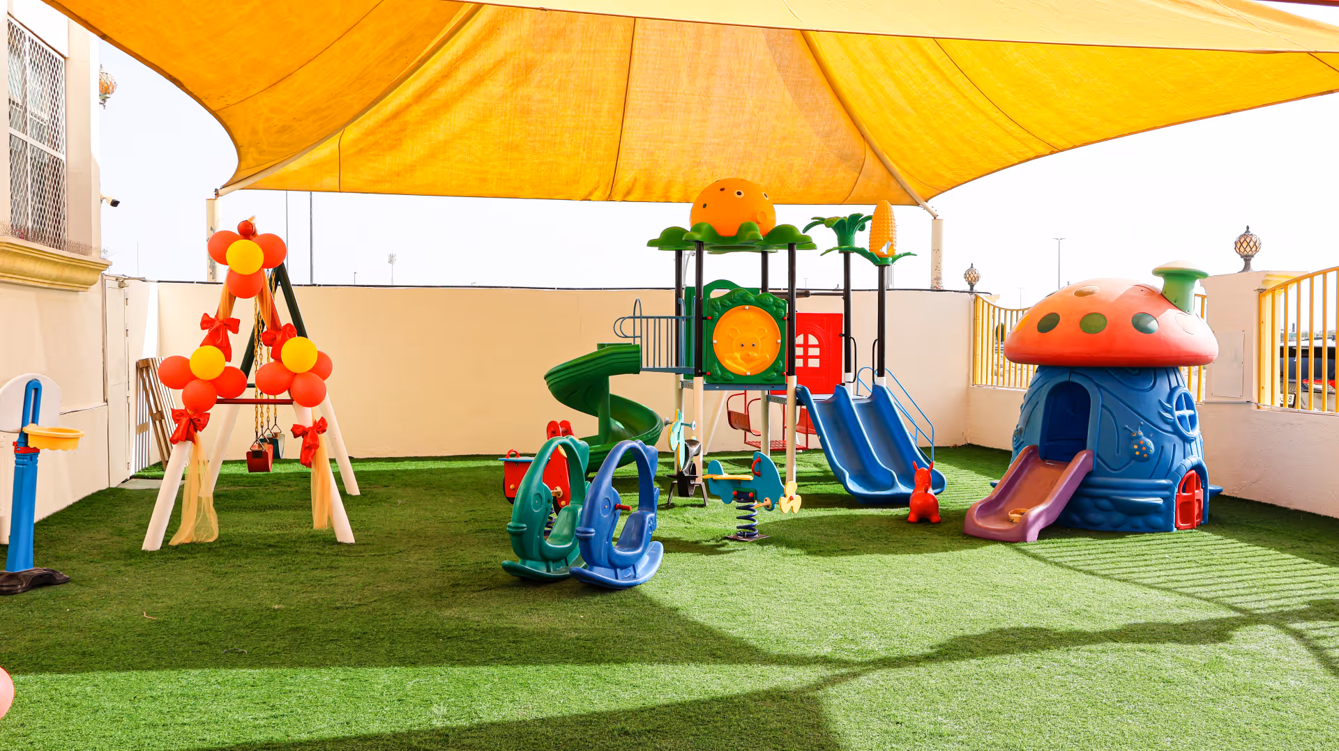 Colorful outdoor playground with swings, slides, spring riders, and a mushroom-shaped playhouse under a large yellow canopy.