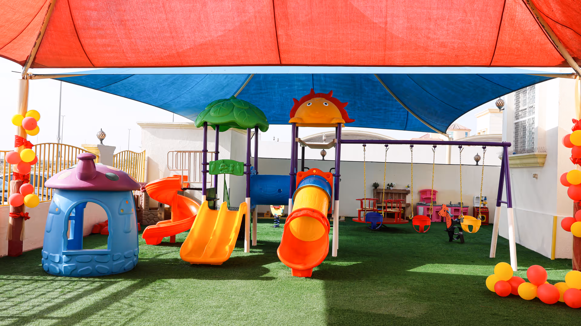 Colorful children's playground with slides, swings, and playhouse under a large blue and red shade.