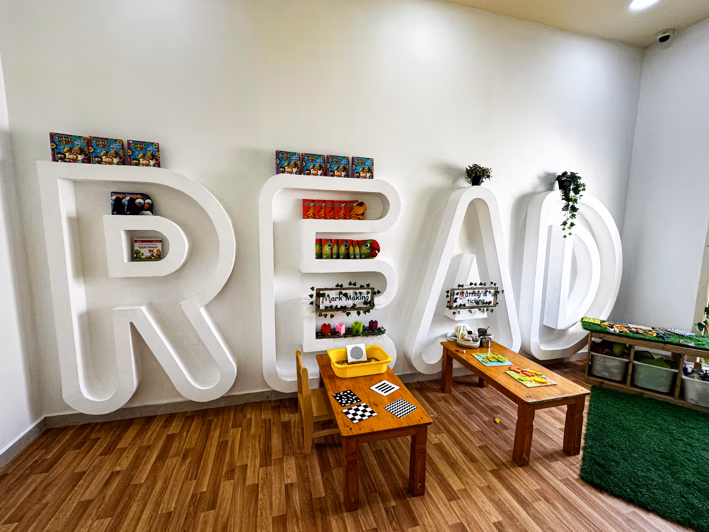 Children's reading area with large white shelves shaped as the word READ holding books and toys, with wooden tables and chairs in front.