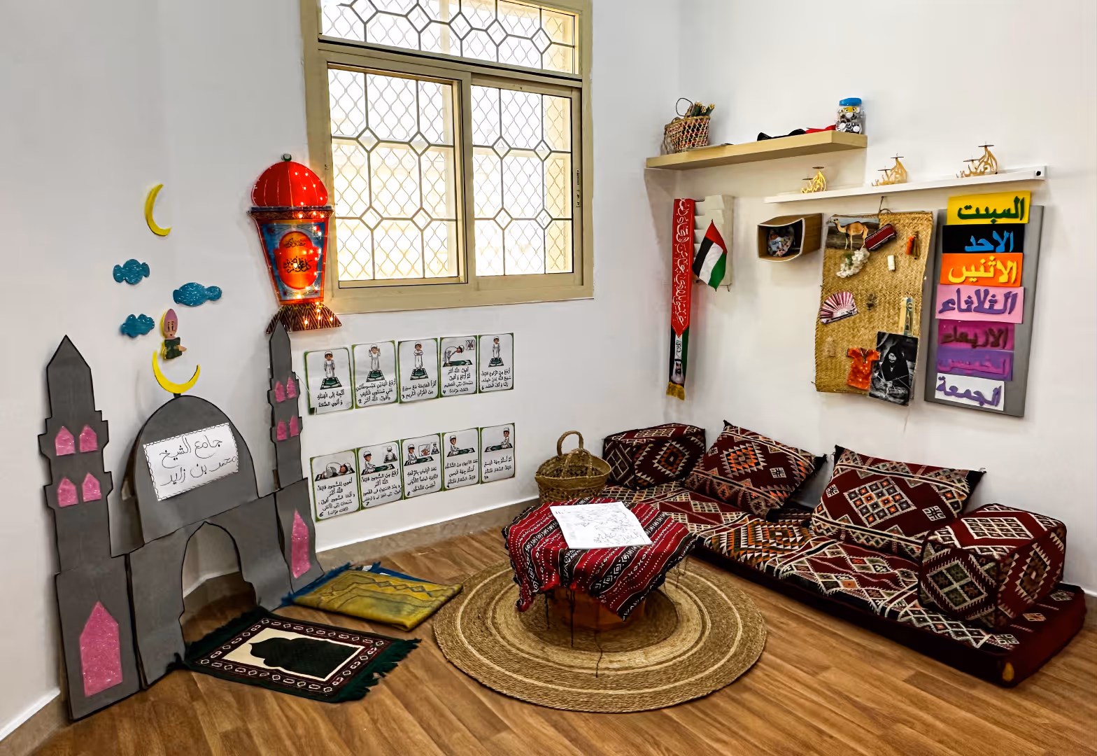 Cozy corner with traditional patterned cushions and seating on floor, woven round rug, a small table covered with a red patterned cloth, wall decorations including Arabic text, a handmade model mosque, and a small shelf.