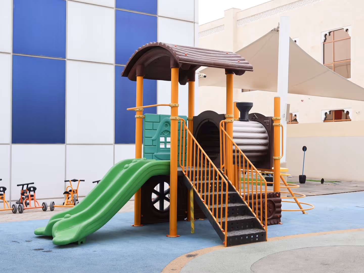 Outdoor children's playground structure with green slides, orange stairs, and a brown roof shaped like a train engine.