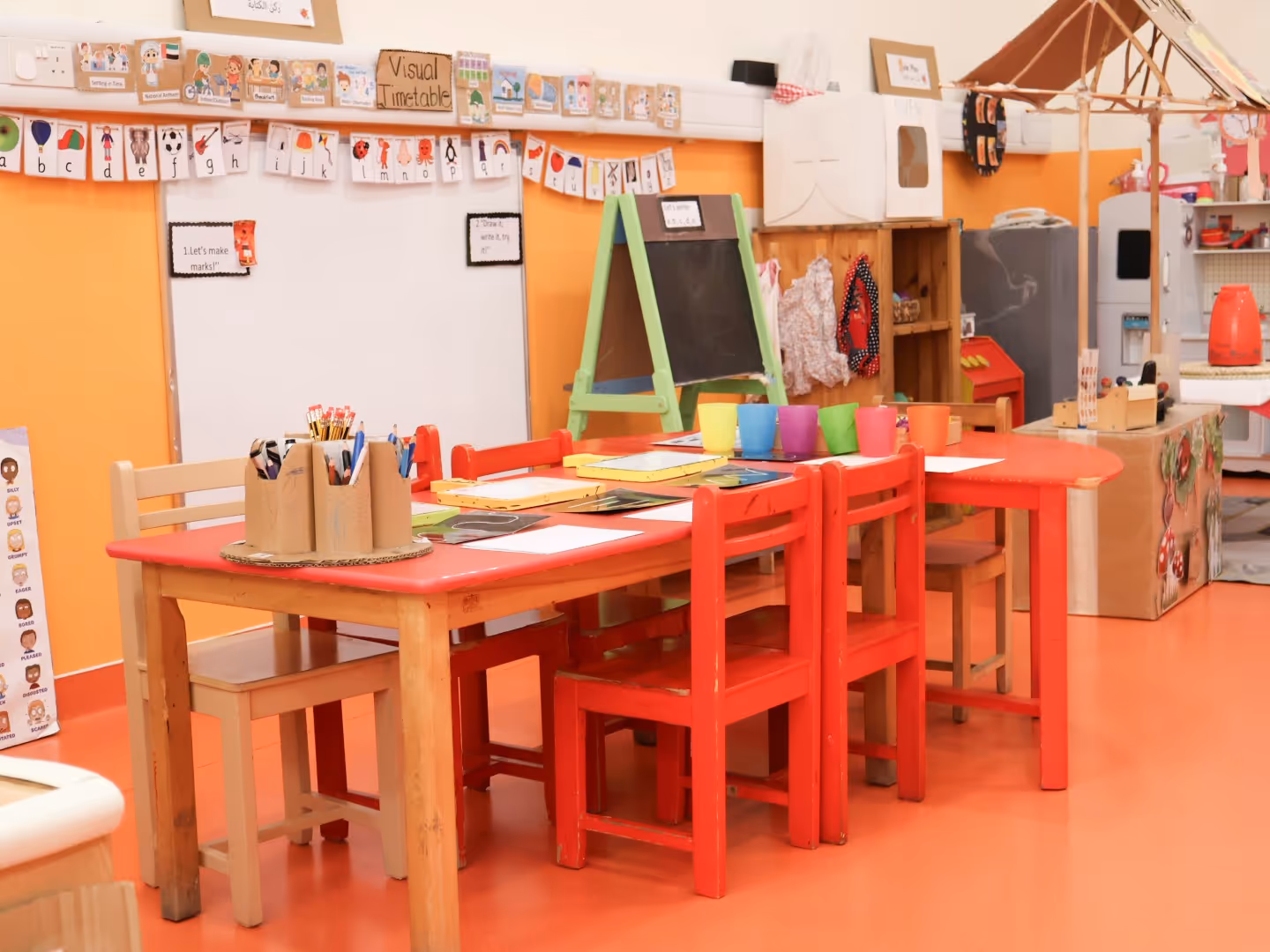 Colorful kindergarten classroom with red and beige chairs around a wooden table, art supplies, and educational posters on the orange walls.