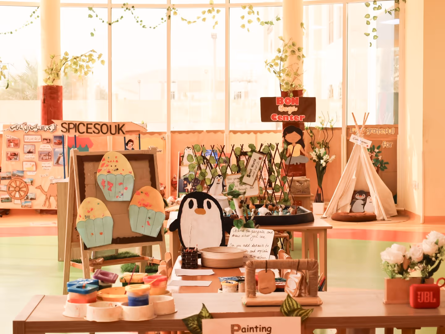 Classroom art and craft display with penguin drawings, cupcakes, plant trellis, play tent, and painting supplies on tables.