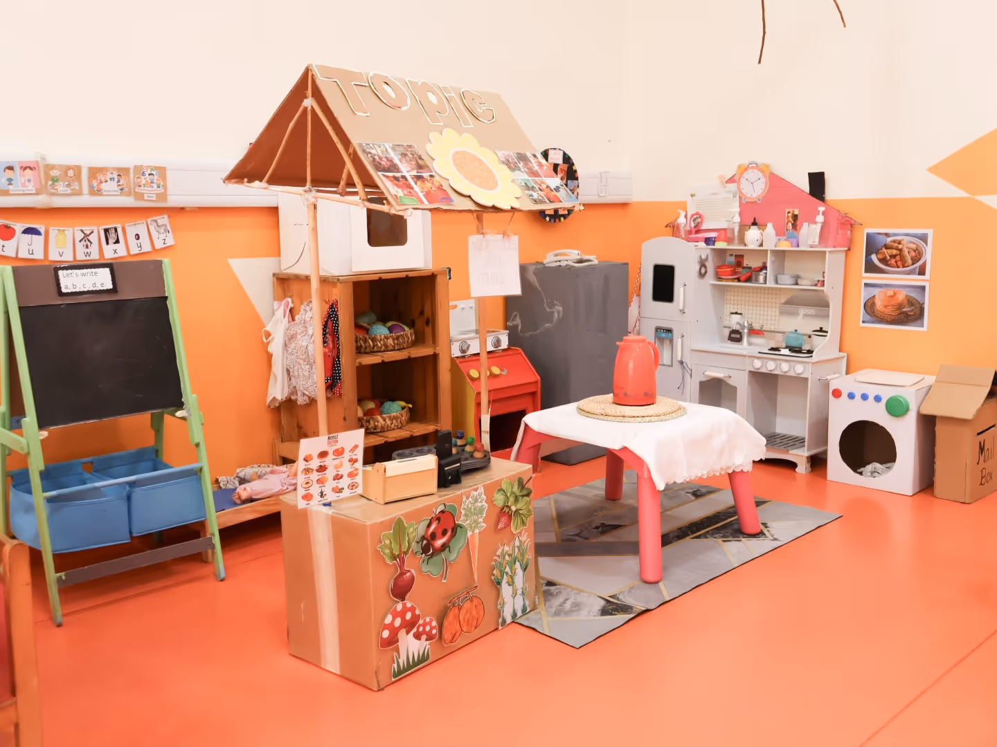 Colorful children's play kitchen area with toy appliances, a table with a kettle, and a small market stall labeled 'TOPIC'.