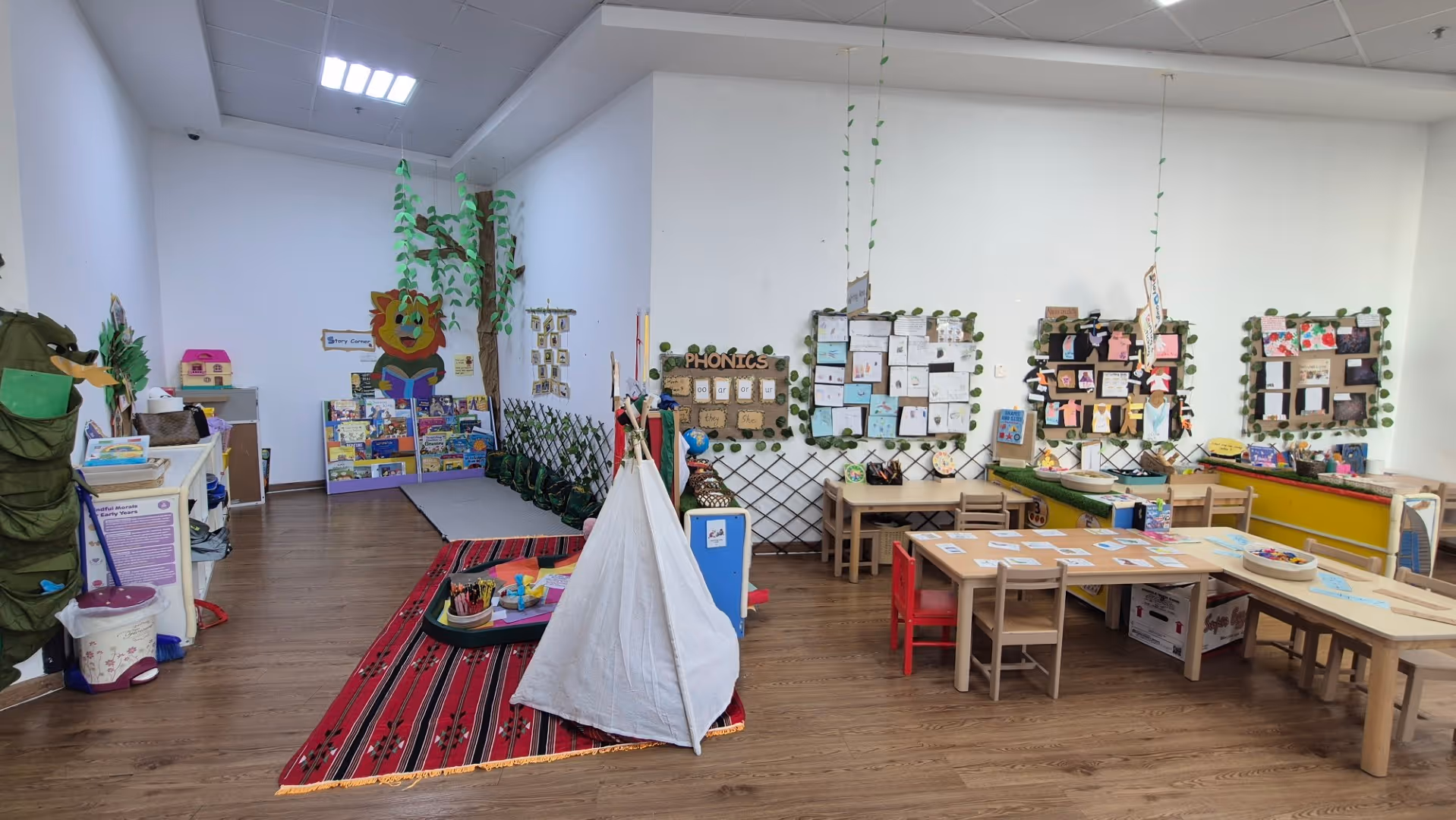 Colorful children's classroom with tables, chairs, bookshelves, a teepee on a red rug, and educational posters on the walls.