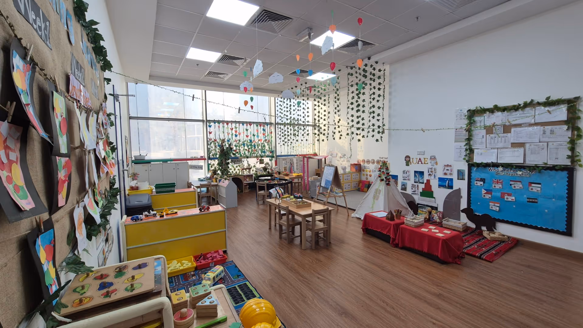 Bright classroom with wooden flooring, children's tables and chairs, colorful artwork on walls, hanging decorations, and various learning centers.