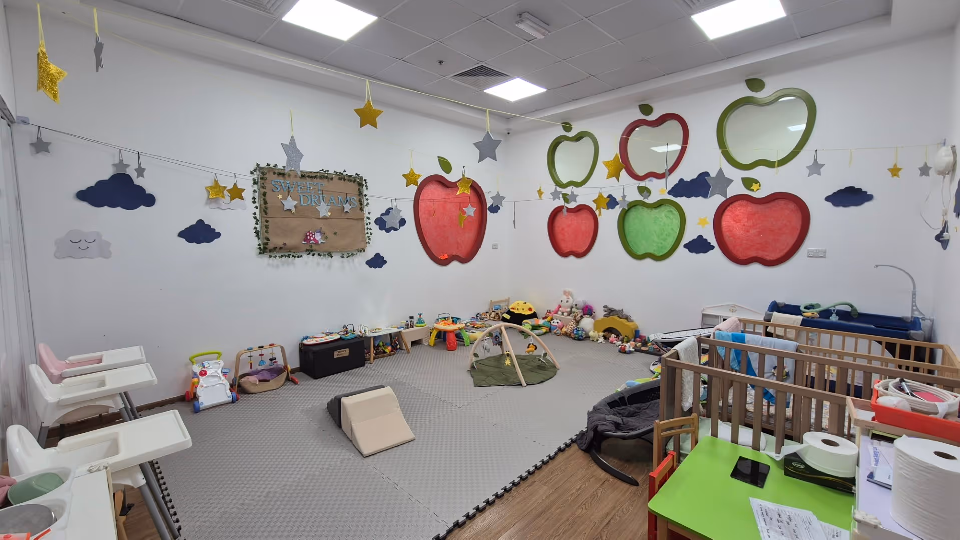 Bright nursery room with play mats, cribs, toys, high chairs, and colorful apple and cloud wall decorations.