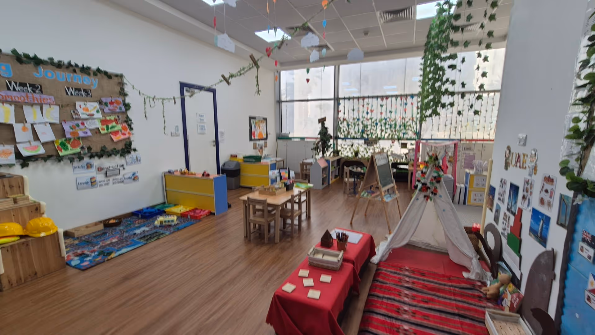 Bright and colorful classroom with wooden floor, small tables and chairs, decorated walls, a play tent, and educational materials.