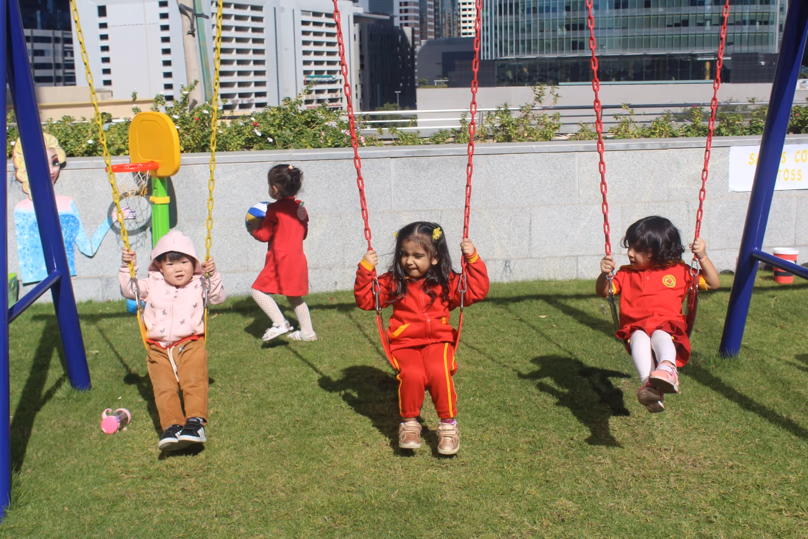Three children sitting on red and yellow swings in a grassy playground with a cityscape background, one child holding a ball.