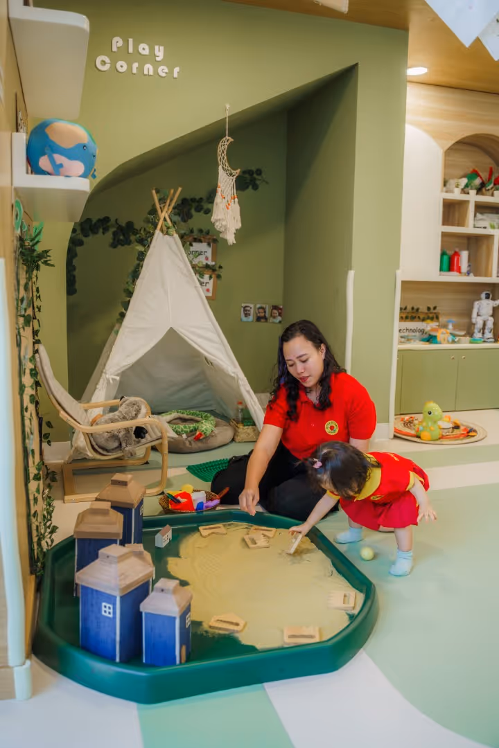 Adult and toddler playing together in a play corner with wooden blocks and a small teepee tent in a cozy, green-themed room.
