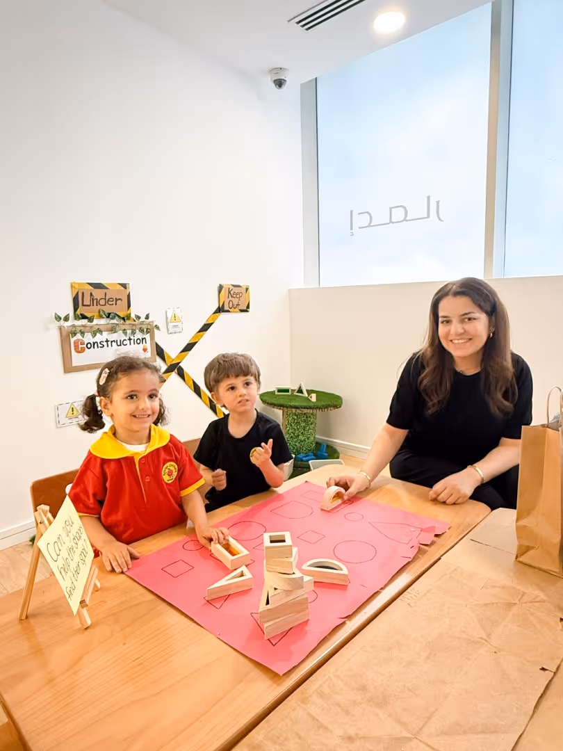 Two children and a woman sitting at a table with pink paper and wooden geometric shapes in a classroom setting with 'Under Construction' signs on the wall.