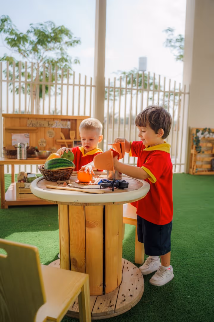 Two young children in red and yellow uniforms playing with toy fruits and vegetables at a round wooden table in a bright indoor play area with green flooring.