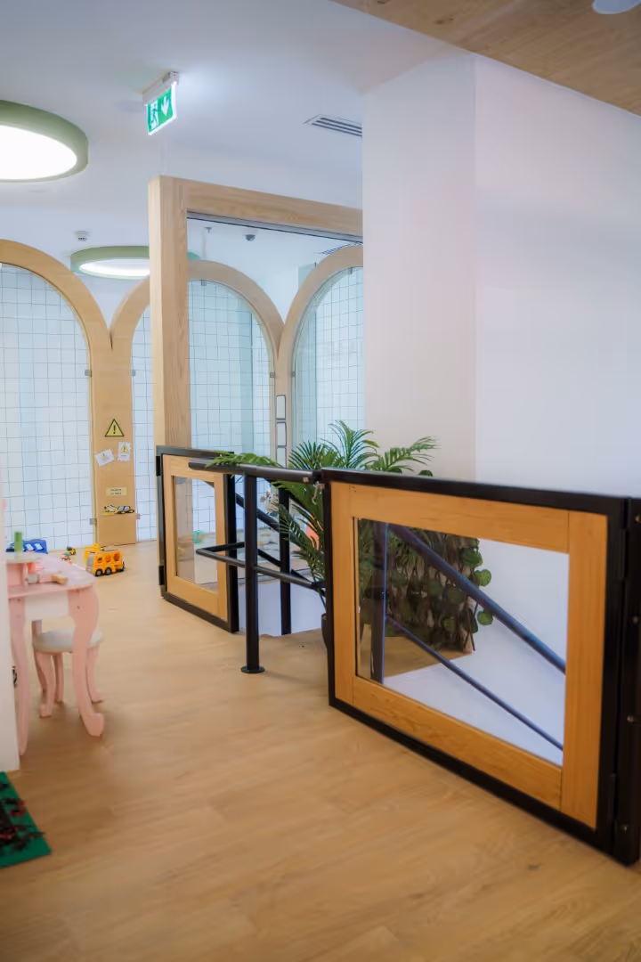 Interior of a modern room with wooden flooring, a small pink table and chair, wooden-framed glass doors, and green plants near a stair railing.