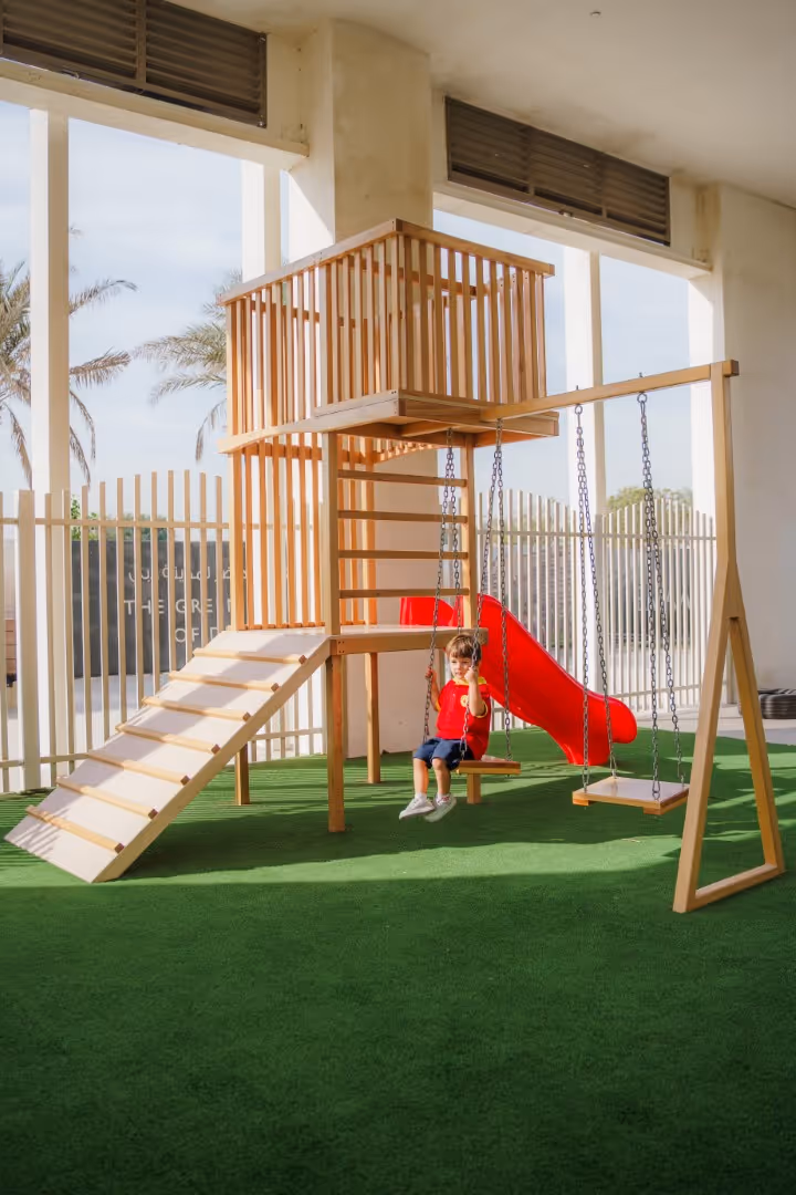 Child in red shirt sitting on a wooden swing set with a slide and climbing ramp on artificial grass under a covered area.