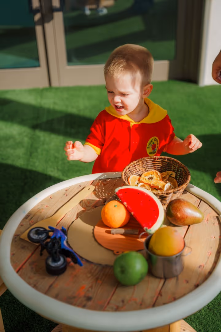 Toddler in red and yellow shirt playing at a wooden table with toy fruits and a basket of dried fruit slices.