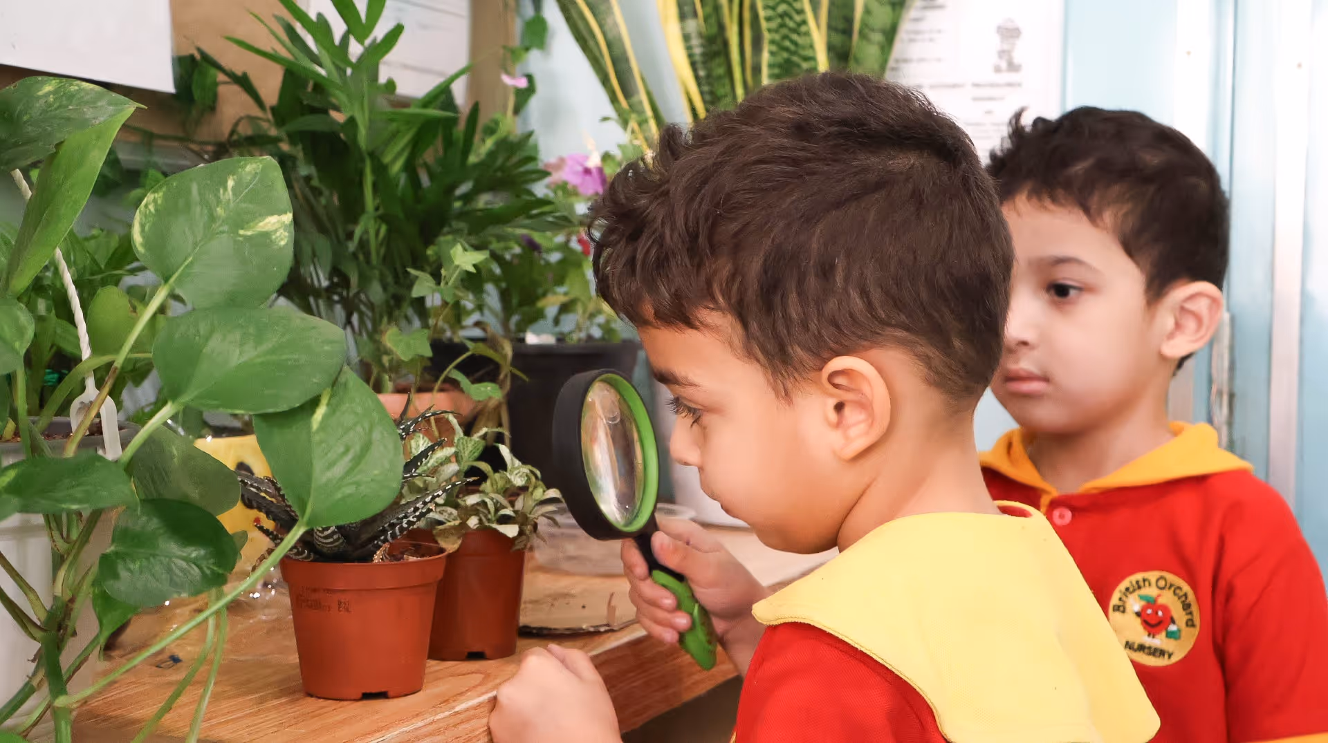 Two young boys in red uniforms examine potted plants closely, one using a green magnifying glass.
