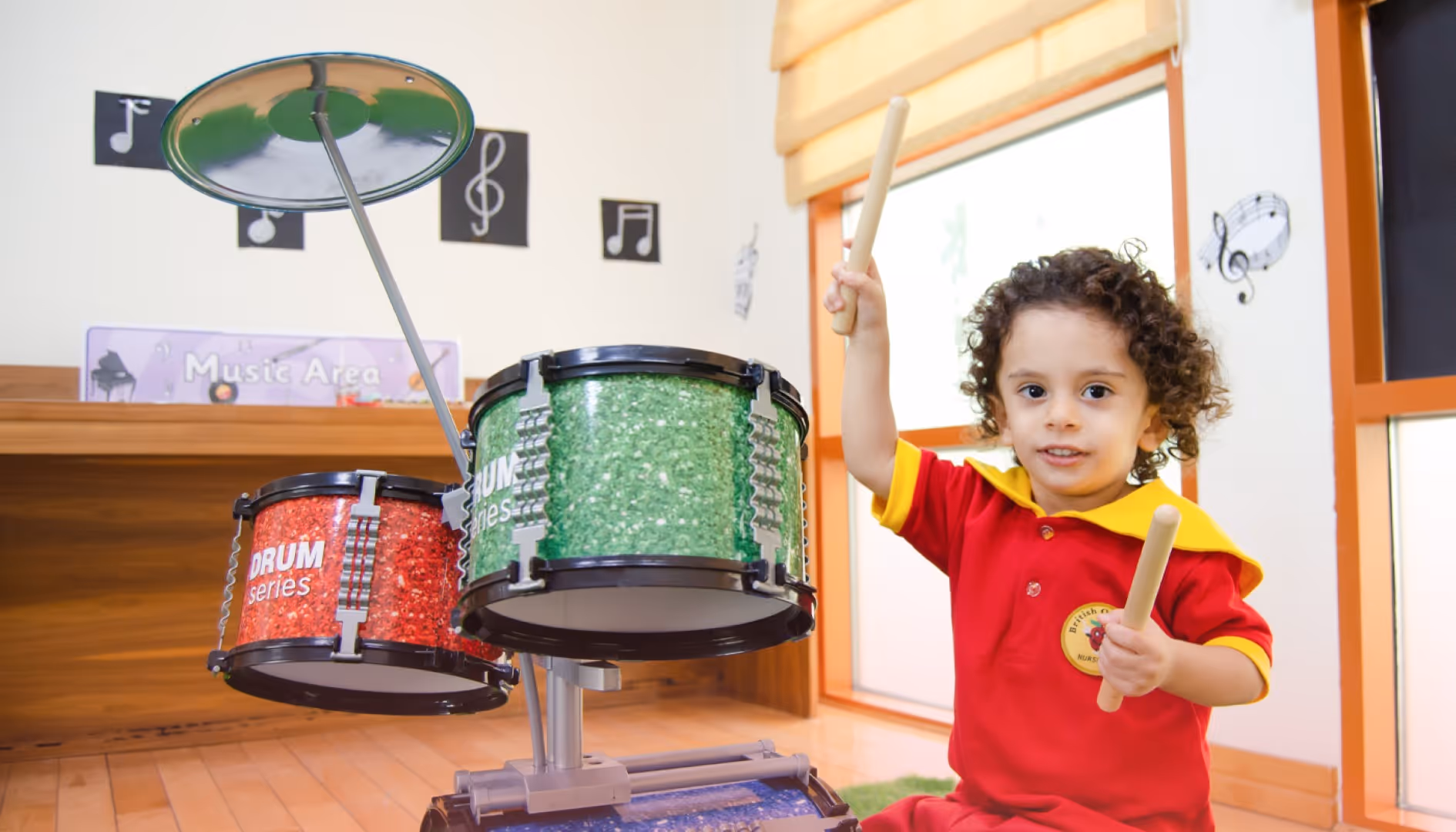 A young child with curly hair in a red and yellow outfit holding drumsticks next to a small colorful drum set indoors.