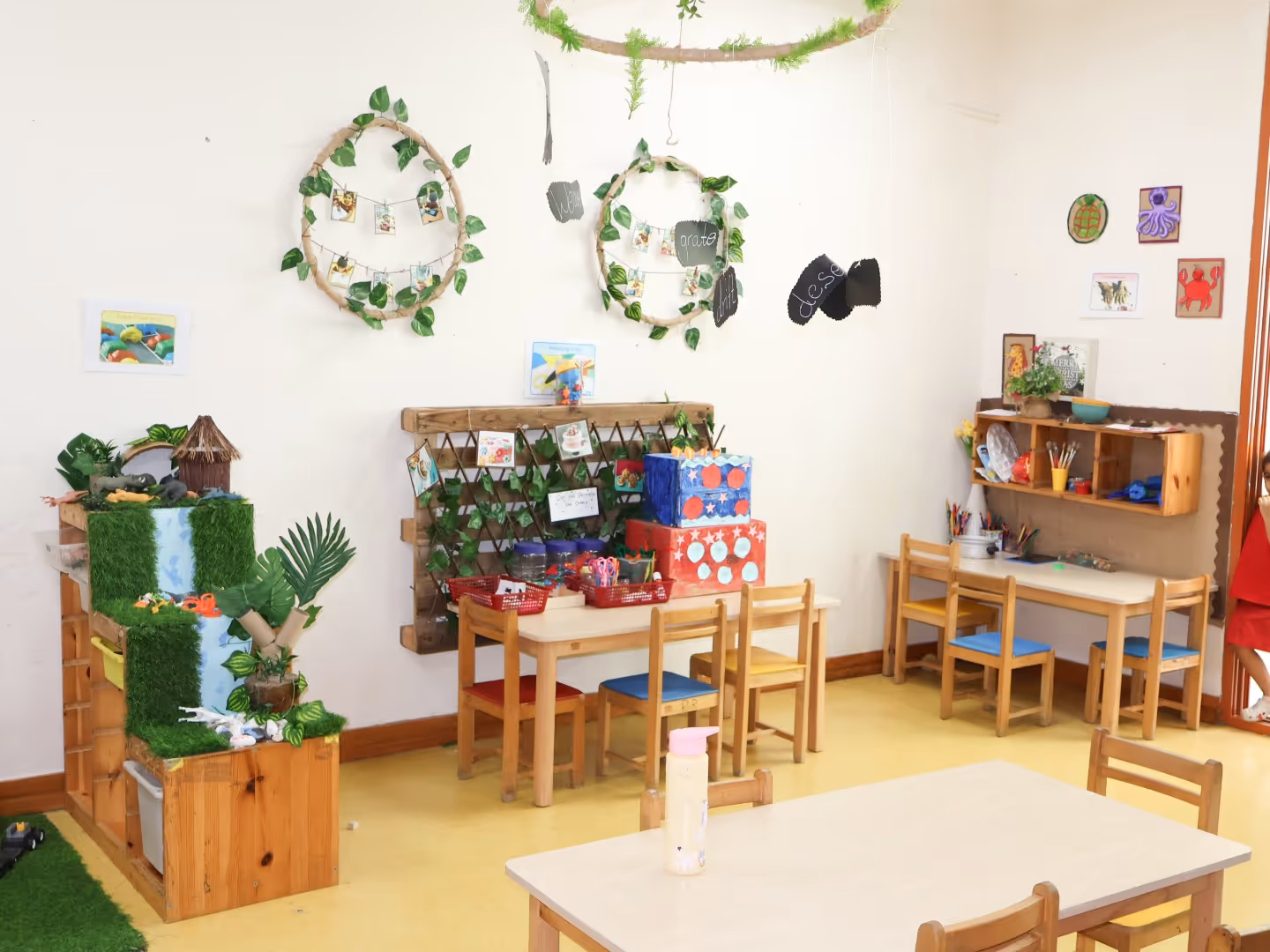 Bright classroom corner with small wooden tables and chairs, decorated with plants, craft supplies, and children's artwork on the walls.