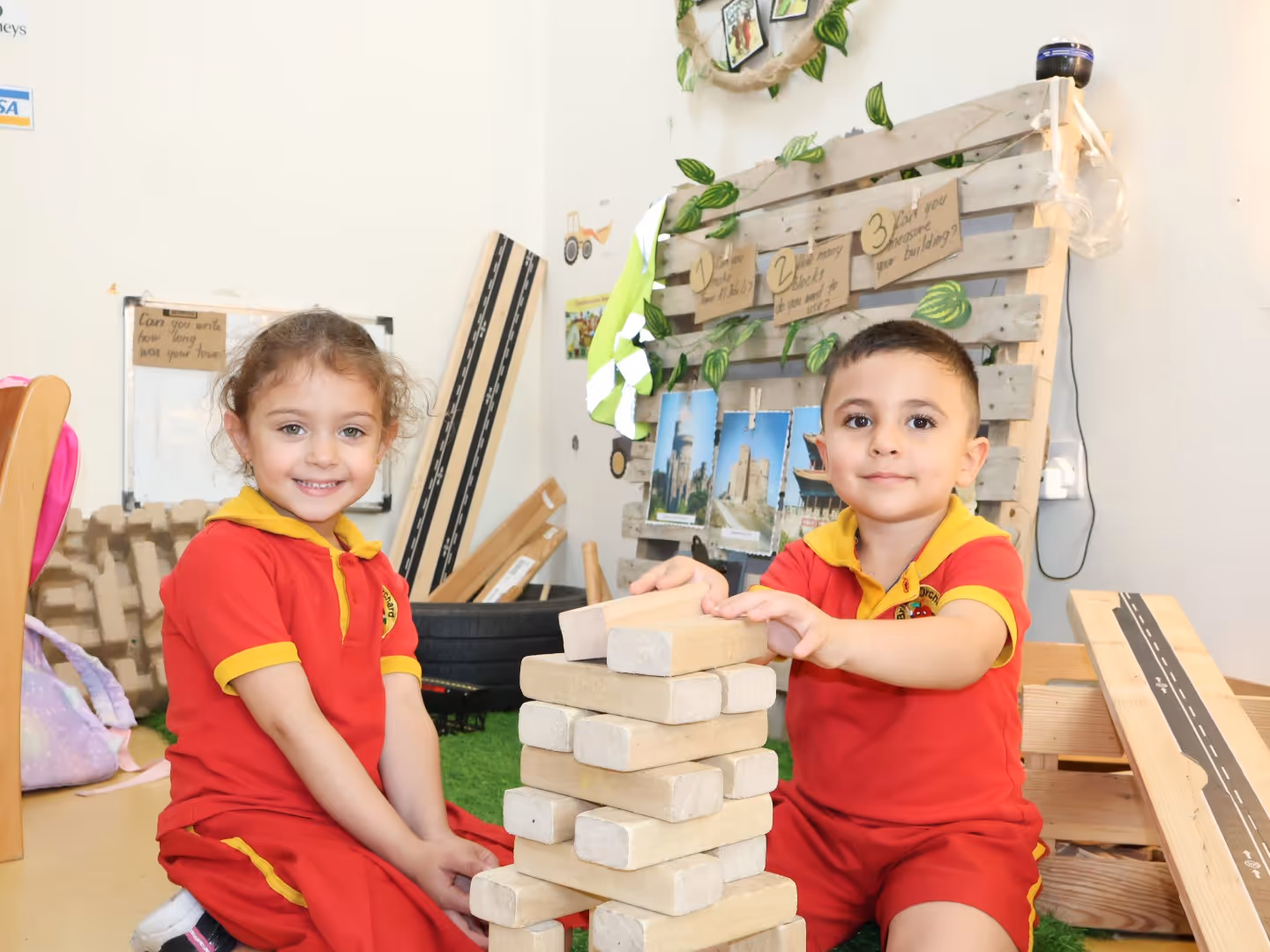 Two children in red and yellow play uniforms stacking large wooden blocks indoors.