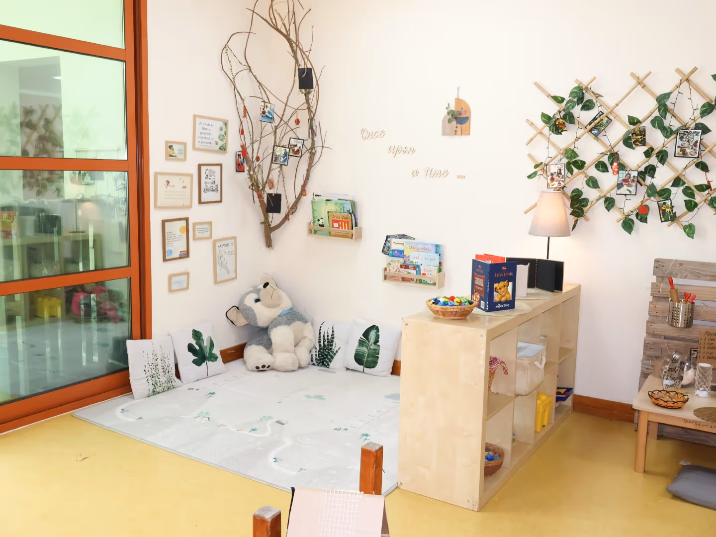Cozy children's reading corner with leaf-patterned pillows, a large bear plush, wall-mounted bookshelves, framed pictures, and decorative vines with photos.