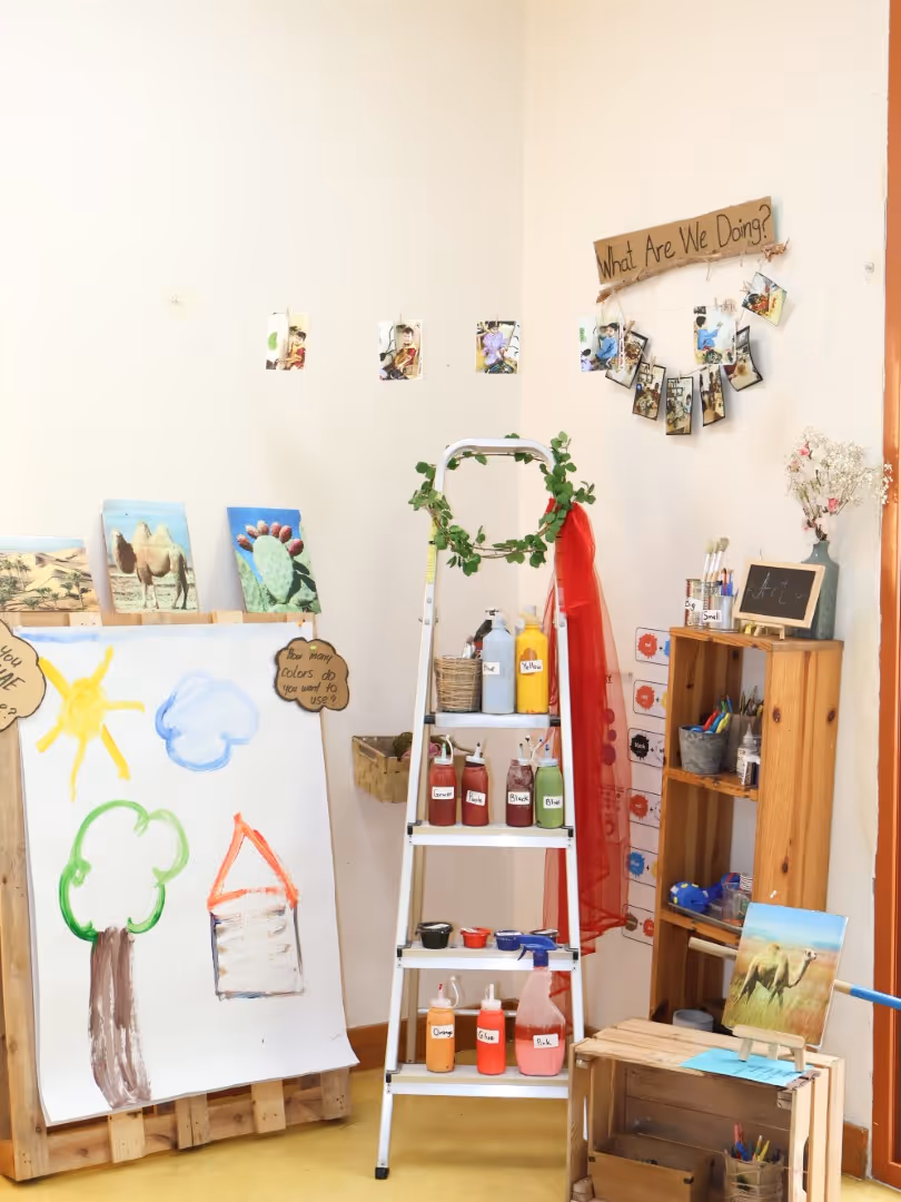 Children's art corner with a large paper showing a sun, cloud, tree, and house, paint bottles on a ladder, and paintings of camels and a cactus.