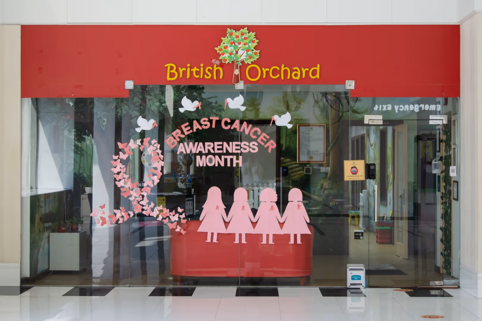 Glass entrance of British Orchard decorated with pink ribbon, silhouettes of four girls holding hands, and text 'Breast Cancer Awareness Month' for breast cancer awareness.
