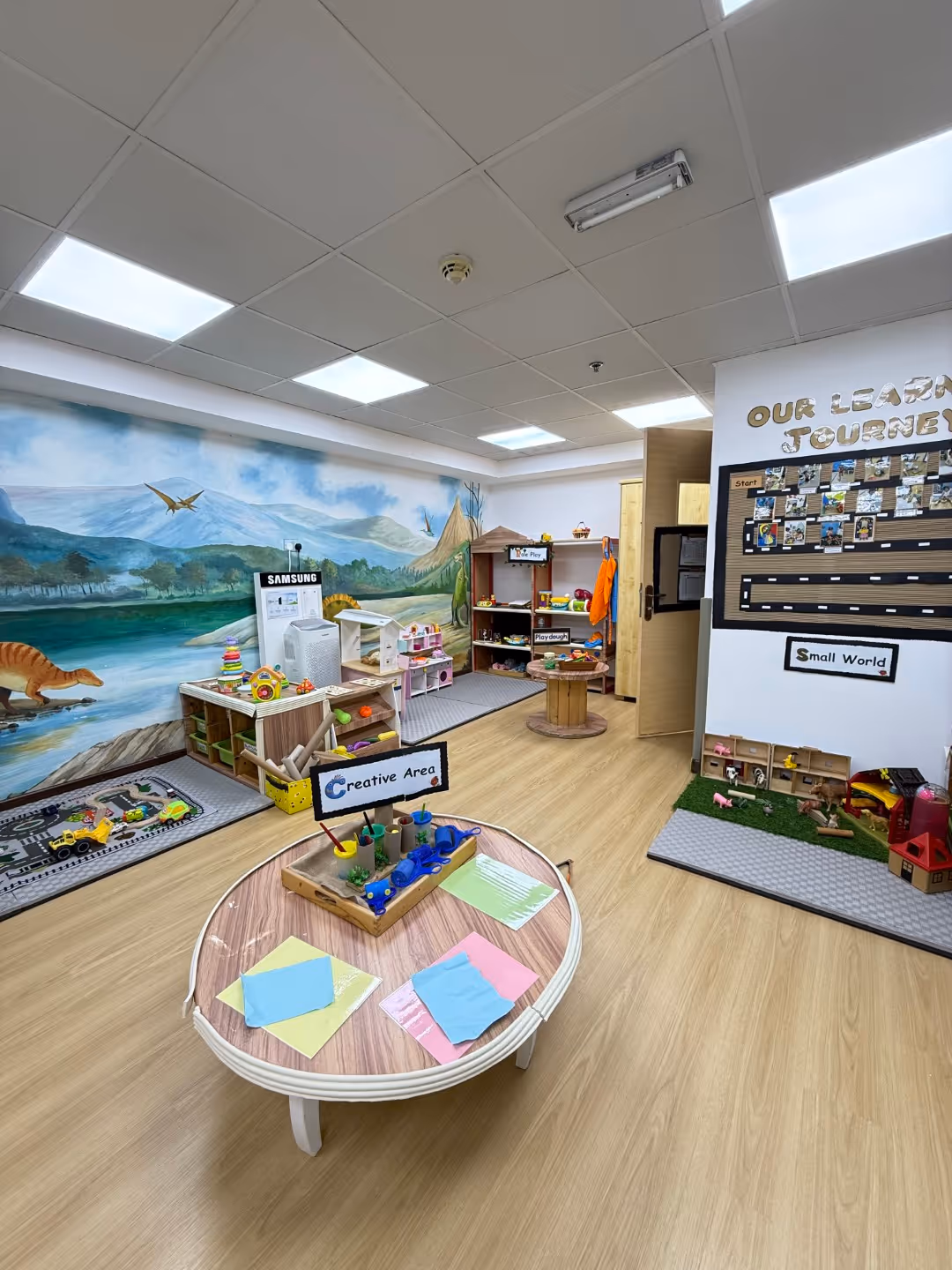 Bright children's playroom with creative and small world activity areas, dinosaur mural, and various toys.